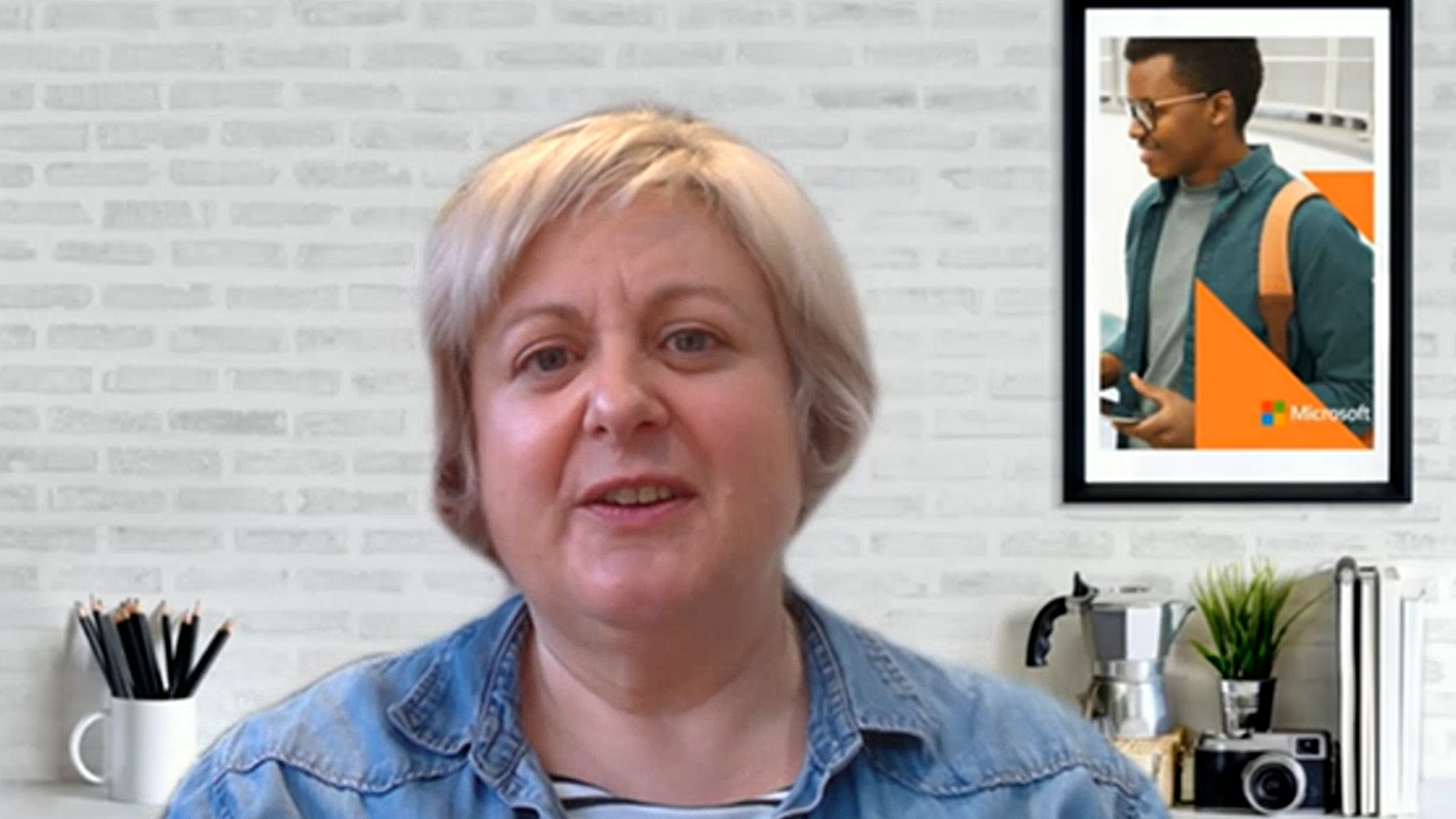 Woman in a denim jacket sitting by a desk with camera, plant, and framed poster.